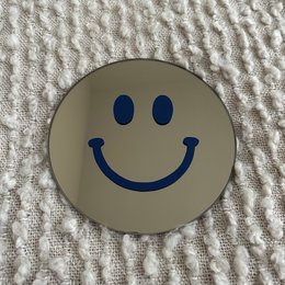A round, dark blue mirror with a smiling face design. The mirror has a simple, minimalist style that would complement a variety of home decor styles.