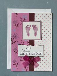 A folding baby card with a pink and brown color scheme, featuring a framed image of baby footprints and the text "Mini-Meisterstück" (German for "masterpiece"). The card has a floral pattern and a ribbon accent.