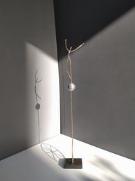 A minimalist metal sculpture with a single hanging pearl-like ornament, standing on a simple base. The sculpture has a sleek, modern design with clean lines and a focus on the interplay of light and shadow.