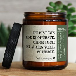 Scented candle in a glass jar with a label that reads "Du bist wie eine Blume, ohne dich ist alles voll scheisse" (You are like a flower, without you everything is full of shit). The candle appears to be a natural vanilla scent.