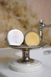 A round, white balm container and a round, yellow balm container on a metal stand against a blurred background.