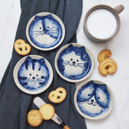 Ceramic cat-themed serving plates in various colors and patterns, including tabby, tortie, calico, and piebald designs. The plates feature whimsical cat faces and are suitable for serving snacks or small bites.