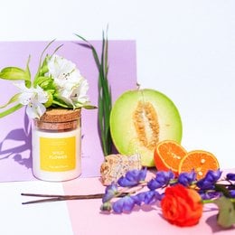 A round, tall orange scented candle in a glass jar with a cork lid. The candle is surrounded by fresh flowers and citrus fruits, creating a vibrant and natural display.