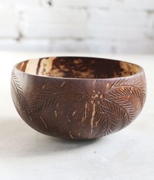 A round, shallow bowl made from a coconut shell with an intricate carved design featuring tropical leaves and patterns.