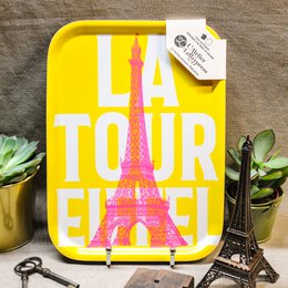 A rectangular yellow tray with the text "La Tour Eiffel" and an image of the Eiffel Tower. The tray is used to hold a cup of tea and some bread on a pink and yellow patterned surface.