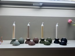 Tall, round candles in various earthy colors including eucalyptus, army green, army brown, and matte black. Displayed on a shelf with abstract ceramic shapes.