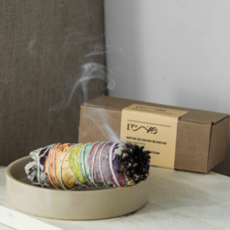 White sage incense stick in a ceramic bowl, emitting smoke. The stick is wrapped in colorful ribbons or fabrics, adding a decorative touch.