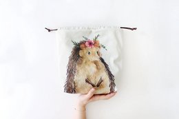  A drawstring bag featuring a cute, whimsical illustration of a hedgehog named Nikki, with a floral crown. The bag is available in three size variants - sock, regular, and sweater.
