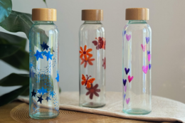 Set of 3 glass water bottles with colorful floral and heart designs. Bottles have wooden lids and are available in various color combinations.