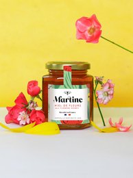  A jar of golden honey with a label that reads "Martine Honey of Flowers". The honey appears thick and viscous, with a few small flower petals visible within.