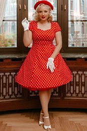 A red polka dot dress with a flirty 1950s style. The dress features a fitted bodice, short sleeves, and a flared skirt. The dress is made of a stretchy, comfortable material and is available in sizes 8 to 26.