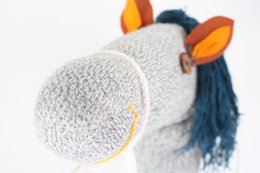  A plush gray horse toy with orange accents and a long mane. The toy appears to be made of soft, textured material and has a wooden stick handle for use as a rocking or riding toy.
