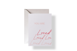 A white greeting card with the text "You are loved" written in red cursive script. The card has a simple, minimalist design.