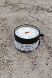 A round, black tin candle with a white wax surface, surrounded by tall green grass. The candle is labeled "Travel tin 90g scented candle - Sunlit Shores".