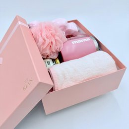 A pink gift box containing wellness and beauty products, including a pink bath bomb, a pink lotion bottle, and a pink fluffy pom-pom.