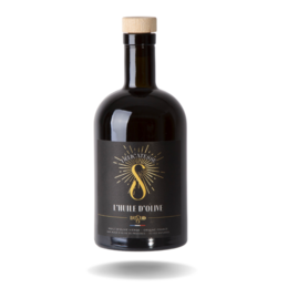 Dark colored olive oil in a glass bottle with a cork stopper. The bottle has a label with a sun design. The oil is accompanied by a loaf of bread on a wooden surface, suggesting it is a high-quality cooking oil from the Provence region.