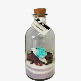A clear glass bottle containing a floral arrangement with a teal flower and dried petals. The bottle has a cork stopper.
