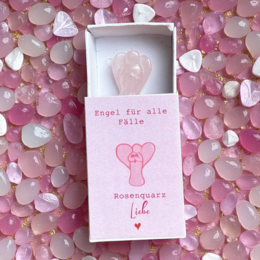  A greeting card with a rose quartz angel figurine and a pink heart design. The card is enclosed in a soft, cream-colored drawstring bag with a pink heart motif.