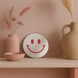 A round, mirrored wall decor with a pink smiley face design. The mirror has a simple, modern style and appears to be made of metal or glass.