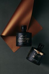 Dark chocolate liqueur in a glass bottle with a dark label. The bottle is surrounded by dark red and brown fabrics, creating a moody, sophisticated atmosphere.