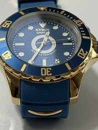 A large, gold-toned watch with a blue dial and bezel. The watch features a date display and a textured, rubber-like strap in a matching blue color.