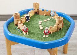  A round, blue plastic tray with a grassy insert and various wooden toy figures, including a castle and people, creating a sensory play scene.