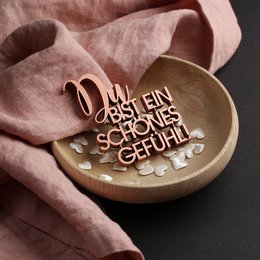 A small, round wooden plaque with the German text "Du bist ein schönes Gefühl" (You are a beautiful feeling) carved into it, surrounded by decorative elements. The plaque is placed on a soft, pink fabric.