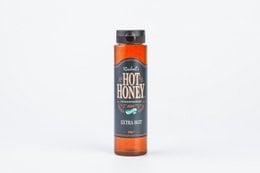 Spicy hot honey sauce in a glass bottle with a black cap. The bottle is orange in color and has the text "Rachel's Hot Honey Extra Hot" printed on it. The sauce is accompanied by some fried chicken or wings on a wooden board.