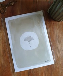 A minimalist poster featuring a simple white ginkgo leaf silhouette against a neutral background. The poster has a Japandi style aesthetic and is printed on sustainable recycled paper.