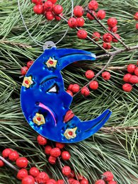 Decorative blue moon-shaped ornament with yellow stars, surrounded by green pine needles and red berries.