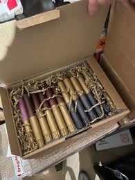 Assorted candles in a variety of colors and shapes, including tall, round, and decorative styles, packaged in a wooden box with shredded paper filler.