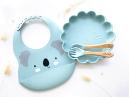 A fun lion-themed mealtime set for kids, featuring a sky blue, pink, yellow, and ocher color palette. The set includes a bib and coordinating plates, bowls, and utensils with a playful lion design.