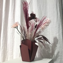 A tall, rectangular paper vase in a burgundy color, filled with a bouquet of dried flowers and foliage in shades of pink, burgundy, and beige.