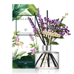 Lavender diffuser with glass container and purple floral arrangement on a table.
The image shows a lavender diffuser with a glass container and a purple floral arrangement on a table. The diffuser appears to be the Cocodor brand, as indicated by the product title. The floral arrangement includes purple flowers, likely lavender, and green leaves. The diffuser and floral arrangement are placed on a white surface, likely a table, with a potted plant visible in the background.