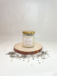 Tall, round lavender-scented candle in a glass jar with a gold lid. The candle is placed on a wooden surface with dried lavender petals scattered around it.