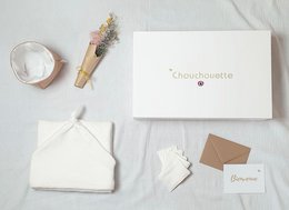 A white gift box with the text "Birth Gift Box 'Mom and Baby'" on the lid, containing a soft white swaddle blanket and a small decorative item.