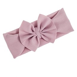 A soft, pink bow headband for babies and young children. The headband features a large, decorative bow made of a ribbed fabric in a light pink color. The headband is available in sizes ranging from 0-6 months up to adult.