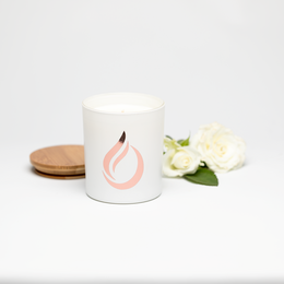 A white soy candle with a pink flame icon, surrounded by white flowers. The candle is part of an aromatherapy collection called 'Bloom'.