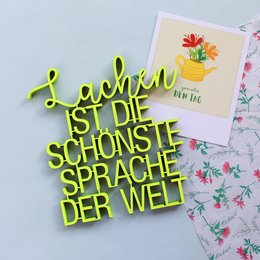A green, cursive-style text that reads "Lachen ist die schönste Sprache der Welt" (Laughter is the most beautiful language in the world) on a light background with a floral postcard.