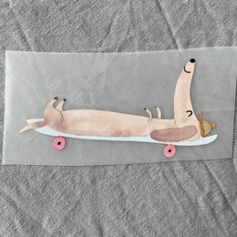 Decorative patch featuring a whimsical dachshund dog design on a skateboard. The patch has a pink and yellow color scheme with a playful, cartoon-like style.