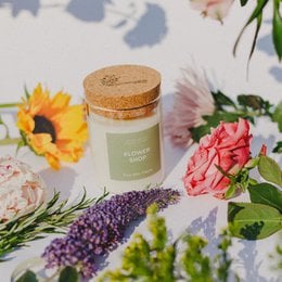 Floral scented candle in a glass jar with a cork lid. The candle is surrounded by fresh flowers, including sunflowers and pink roses. The candle is labeled "Flower Shop" and comes in various sizes.