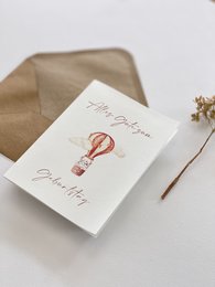 A white greeting card with a hand-drawn hot air balloon illustration and the text "Happy Birthday" written in a cursive font. The card is accompanied by a kraft paper envelope.