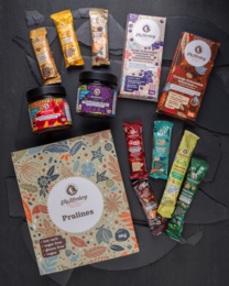 This deluxe gift box from Elly Momberg contains an assortment of sugar-free, vegan, and gluten-free chocolate products. The box features a floral design with various shapes and patterns, including leaves, flowers, and abstract elements. The products inside the box appear to be a variety of chocolate bars and other confections.