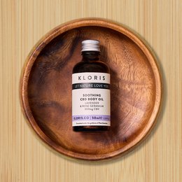 Soothing CBD body and massage oil in a glass bottle with a wooden tray. The oil is amber-colored and the bottle has a white label with the brand name "Kloris" printed on it.