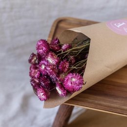 Dried purple helichrysum flowers wrapped in brown paper, with a pink sticker on the package. The flowers have a delicate, wispy appearance and a vibrant purple color.
