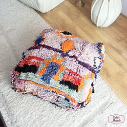 A round, plush Moroccan-style pouf in shades of pink and beige with a geometric pattern, suitable for use as an ottoman or extra seating.
