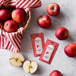 Red apples in a woven basket, accompanied by red and white striped fabric and apple juice cartons. The apples appear to be fresh and ripe, with a vibrant red color.