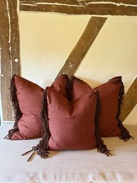 Decorative cushion with burgundy red color, square shape, and fringe trim. Suitable for living room or bedroom decor.