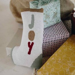A white and red stocking-shaped cushion with a "Joy" text design, placed on a yellow and patterned fabric background near a Christmas tree.