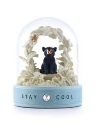 A snow globe featuring a black bear figurine sitting on a blue base with the text "STAY COOL". The globe is surrounded by a snowy, winter scene with white leaves or branches.